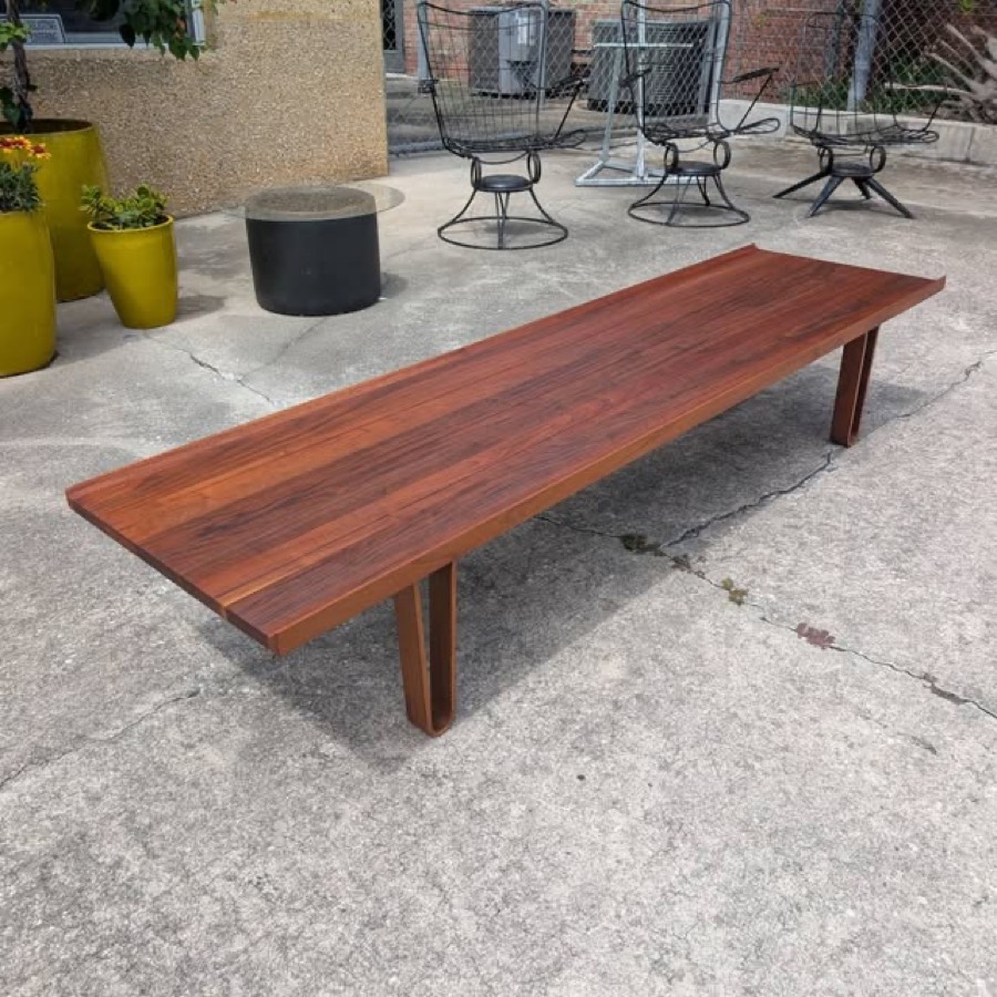A long rosewood slab bench on two plank legs, photographed on weathered grey concrete, with stacked chairs and yellow-green planters behind it.
