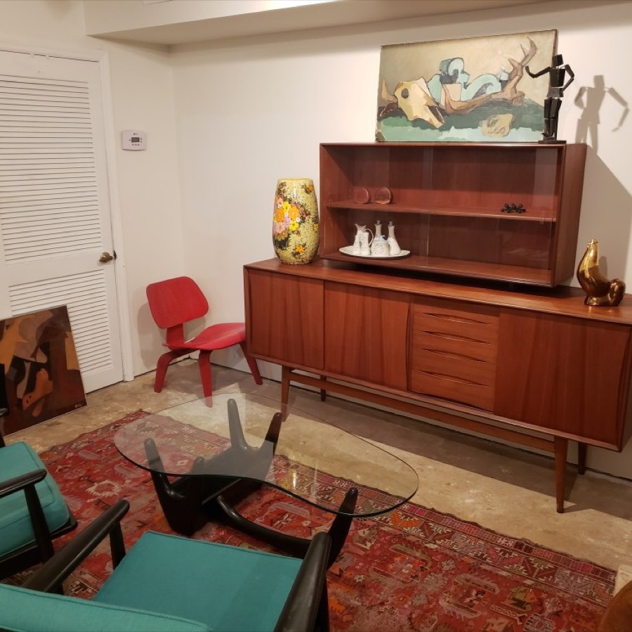 A teak sideboard with a glass-fronted hutch against a white wall, a red moulded plywood chair beside it and a patterned rug on the floor.