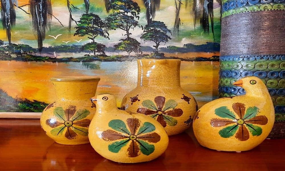 Five mustard-yellow glazed earthenware vessels, two of them shaped like birds, standing on a polished wooden table in front of a painted marsh landscape.