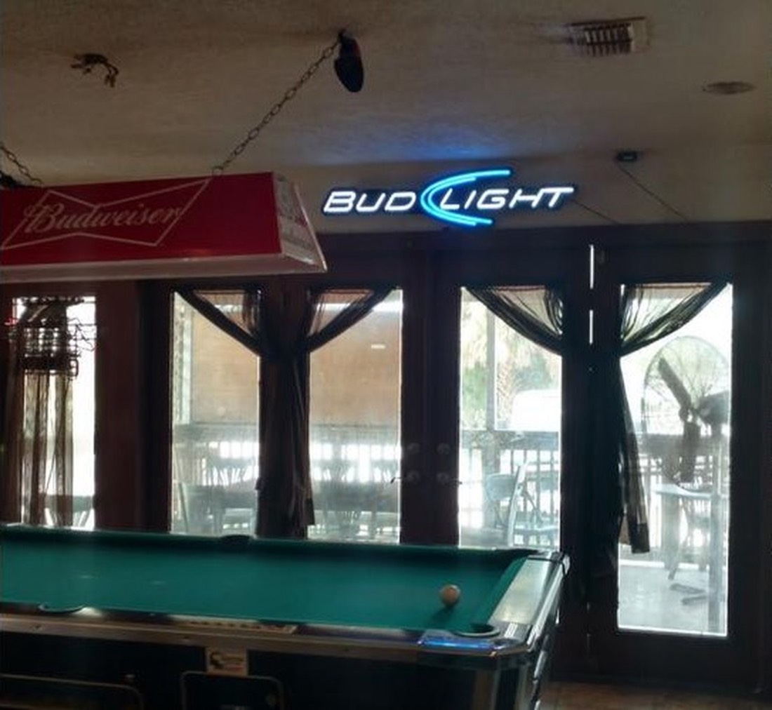 A billiard table with dark green felt in a dim bar, lit from tall curtained windows, with illuminated beer signs above it.