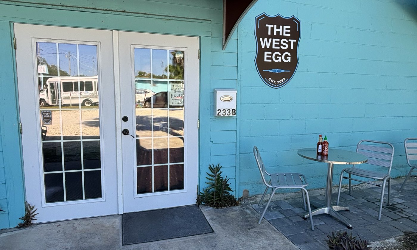 A cinder-block wall painted pale turquoise in full sun, with a white glazed door, a small metal table and two chairs standing on a concrete slab.
